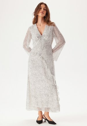 Lange, transparante witte jurk met zwarte spikkels, V-hals en bell sleeves. De zoom heeft een gerimpelde rand, in combinatie met zwarte puntige schoenen.