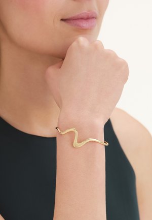Woman with light skin wearing a wavy gold bracelet on her wrist, hand resting near her chin, dressed in a black sleeveless top.