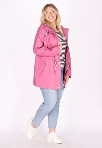 Giacca impermeabile rosa con cappuccio, dotata di tasche frontali, vita regolabile con cordino e dettagli in metallo argentato. Abbinata a jeans azzurri chiari e sneakers bianche.