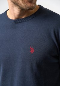Man som bär en marinblå tröja med en liten röd broderad polo-spelarlogo på bröstet, synlig från halsen till midjan.