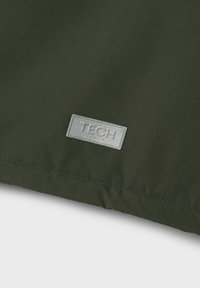 Tessuto verde scuro con una superficie liscia, caratterizzato da un'etichetta rettangolare grigia in rilievo con la scritta "TECH" cucita sul bordo.