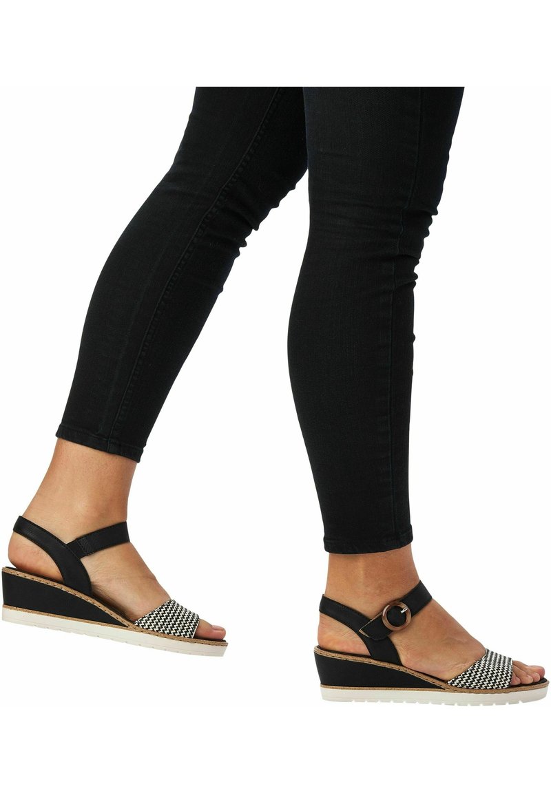Jambes en jean skinny noir portant des sandales compensées noires et blanches avec des brides à la cheville et une boucle, marchant sur un fond blanc.