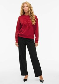 Sweatshirt rouge avec le texte "SOIS TOI" en une teinte de rouge plus foncée, associé à un pantalon noir à jambes larges et des chaussures noires à talons pointus. Texture du tissu lisse.