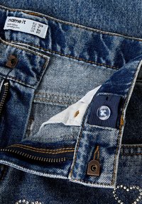 Džinsai iš mėlynos medžiagos su kontrastiniu oranžiniu siuvimu, užtrauktuku priekyje, sidabriniais metaliniais elementais ir audiniu žyma su dydžio informacija.