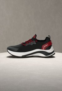 Baskets de sport noires avec lacets rouges et accent au talon, semelle épaisse blanche et noire, présentées sur une surface claire avec un fond gris.