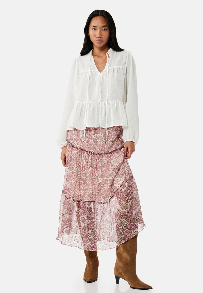 Vrouw draagt een witte blouse met lange mouwen, een roze midi-rok met patroon en lagen, en bruine suède laarzen met hak, staand tegen een effen achtergrond.