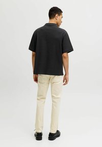 Homme debout, de dos, portant un polo noir à manches courtes, un pantalon beige et des chaussures noires, sur un fond clair uni.