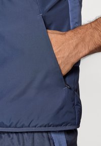 Veste de sport bleu marine avec une texture lisse, dotée d'une poche latérale et de détails contrastants bleu clair le long des coutures.