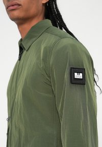 Groen lichtgewicht shirt met een gestructureerde afwerking, een opstaand kraagje en een zwarte patch met witte letters op de linkermouw.
