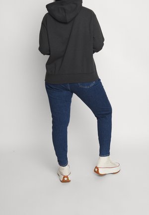 Osoba w czarnej bluzie z kapturem, niebieskich obcisłych dżinsach i białych platformowych trampkach z gwiazdkowym wzorem, stoi na tle jasnego, gładkiego tła.