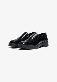 Melni slip-on apavi, kas izgatavoti no spīguļojošas ādas, ar noapaļotu purngalu un biezu, cietu gumijas zoli. Augšdaļā ir smalki šuvju akcenti.