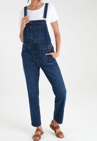 Salopette en jean avec bretelles réglables, poche avant et coupe décontractée. Couleur bleu foncé, portée avec une chemise blanche et des sandales marron.