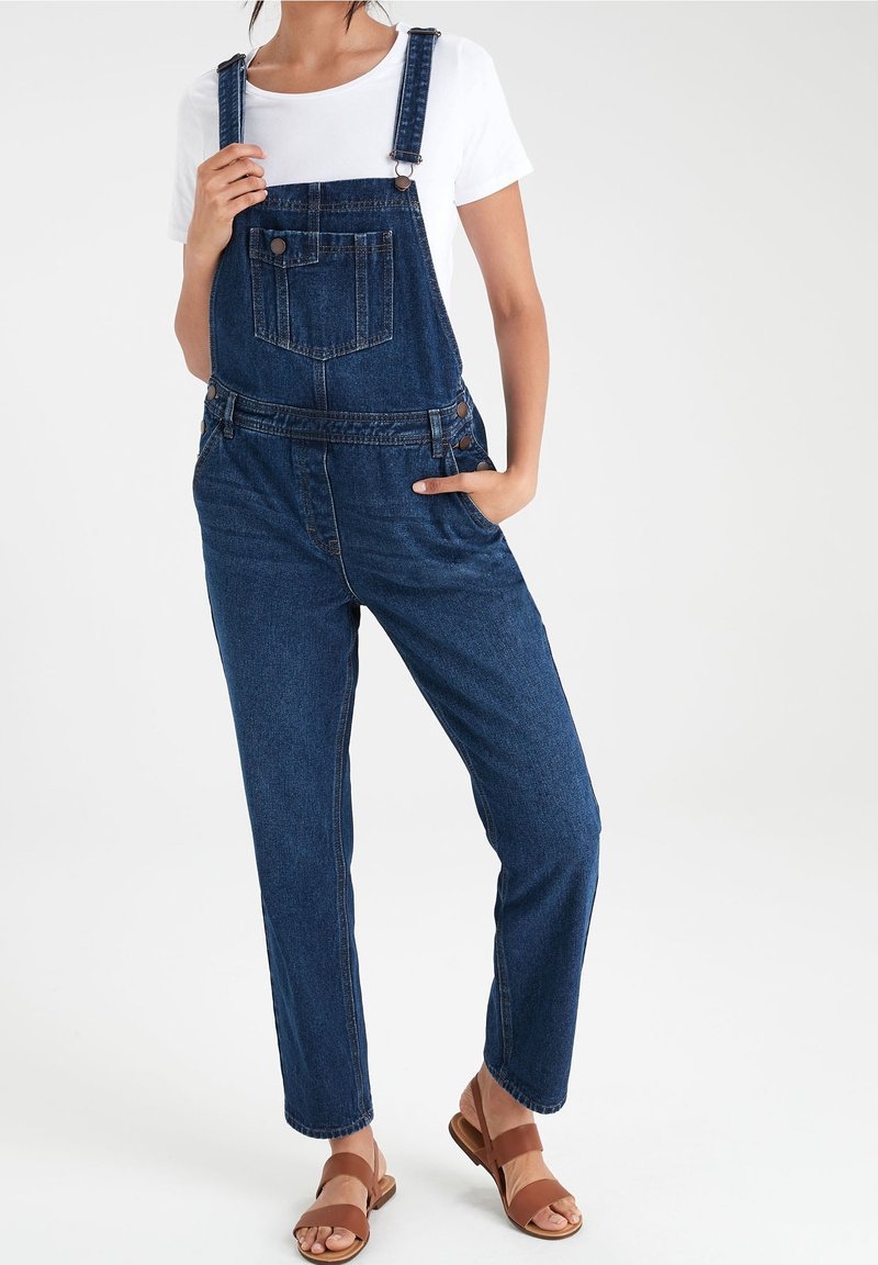 Salopette en jean avec bretelles réglables, poche avant et coupe décontractée. Couleur bleu foncé, portée avec une chemise blanche et des sandales marron.
