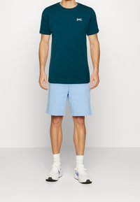 Tyrkysové bavlněné tričko s krátkým rukávem a logem nůžek, spárované se světle modrými šortkami, bílými sportovními botami a bílými ponožkami.