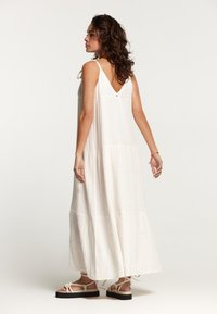 Robe maxi blanche avec des bretelles fines, un dos en V et un design à volants. Fabriquée en tissu texturé. Portée avec des sandales nues à lanières.