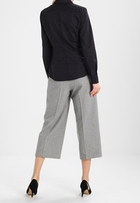 Camicia nera aderente con maniche lunghe, abbinata a pantaloni grigi a gamba larga e tacchi neri, che mettono in risalto un look sartoriale e texture morbide.