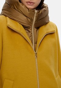 Cappotto di lana giallo con colletto alto, presenta una giacca imbottita marrone sotto, chiusura con zip e tasche laterali. Texture liscia con un aspetto caldo.