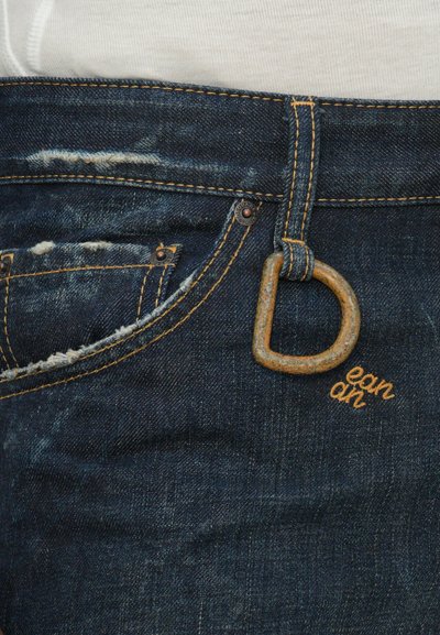 Dark blue denim jeans with distressed details. Features brass hardware, a large rust-colored D-ring, and orange stitching.