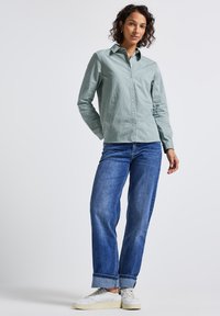 Vrouw met krullend haar die een groen gestreept overhemd met knopen, blauwe opgerolde jeans en witte sneakers draagt, staande tegen een effen achtergrond.