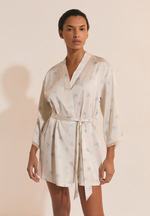 Donna che indossa un accappatoio corto di colore beige chiaro con piccoli motivi floreali azzurri, legato in vita, in piedi davanti a uno sfondo semplice.