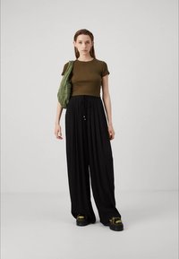 Haut vert ajusté associé à un large pantalon noir, avec une taille à cordon. Le mannequin porte des chaussures noires volumineuses avec des accents dorés.