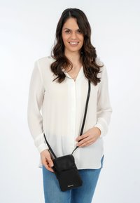 Borsa a tracolla in pelle nera con chiusura a zip e tasca frontale, indossata sopra una blusa bianca con maniche lunghe e chiusura a bottoni.
