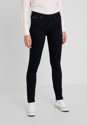 Personne portant un jean slim noir, des baskets montantes blanches et un haut à manches longues rose clair, debout devant un fond uni.
