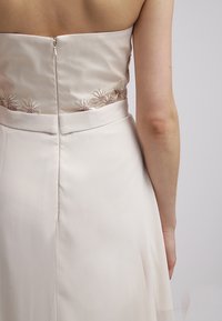 Olkaimeton beige-mekko, jossa on tiukka bodice ja kukkakirjonta. Silkkinen kangas ja vetoketjukiinnitys selässä, laskostettu hameosa.