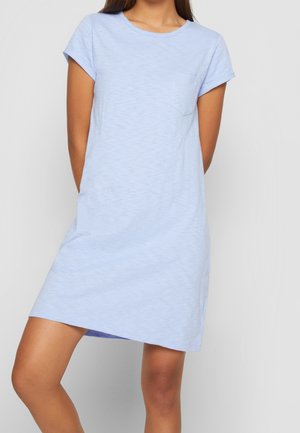 Nuori nainen, joka pukeutunut lyhythihaiseen, vaaleansiniseen rentoon mekkoon, jossa on rintatasku, seisoo yksiväristä vaaleaa taustaa vasten.