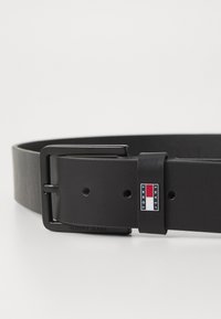 Zwarte leren riem met een matte afwerking, rechthoekige zwarte metalen gesp en een klein embleem met rode en blauwe accenten.