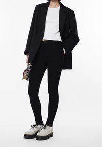 Jeans negros ajustados combinados con una blusa corta blanca y una blazer negra. Zapatos blancos simples con suelas negras. Bolso colorido en la mano.