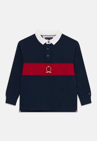 Marinblå långärmad polo med vit krage, röd horisontell rand och guldemblem. Har en tre-knappsknäppning. Tillverkad av bomull.