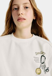 Sweat-shirt blanc avec un logo "Smiley" brodé et un graphique de porte-clés représentant un visage souriant, accentué par du texte en noir et jaune.