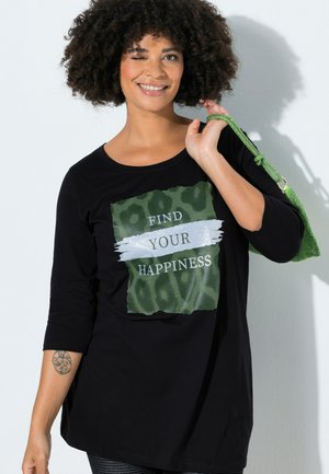 Lächelnde Frau zwinkert, trägt ein schwarzes Shirt mit der Aufschrift "FIND YOUR HAPPINESS" und hält eine grüne Handtasche über der Schulter.