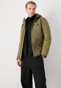 Veste matelassée couleur vert olive avec capuche, dotée d'une fermeture éclair, d'un motif diagonal et de poches latérales. Portée par-dessus une chemise noire à boutons.