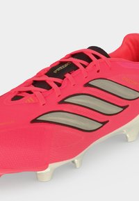 Φωτεινό κόκκινο ποδοσφαιρικό παπούτσι Adidas Predator με μαύρα κορδόνια, τρεις ασημένιες ρίγες στο πλάι και λευκές τάπες στη σόλα.