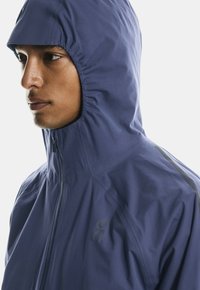 Giacca con cappuccio navy con chiusura a zip, realizzata in un tessuto leggermente strutturato e con dettagli riflettenti sulle spalle. Logo sul lato sinistro.