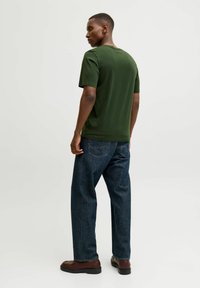 Camiseta verde de manga corta hecha de tela suave, combinada con jeans ajustados de color azul oscuro y zapatos marrones, vista desde la parte de atrás.