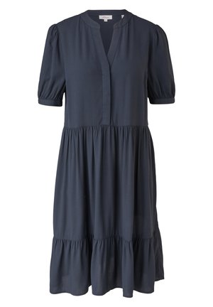 Femme portant une robe bleu marine fluide à manches courtes avec une jupe à volants et des baskets blanches, debout les mains dans les poches sur un fond neutre.