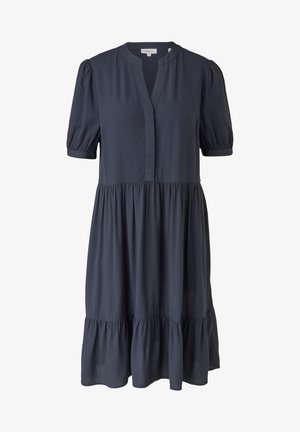 Kvinde iført en løs, kortærmet navy kjole med flæset nederdel og hvide sneakers, stående med hænderne i lommerne på en neutral baggrund.