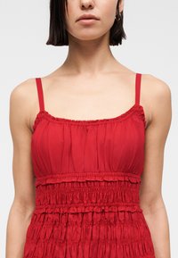 Γυναίκα που φοράει ένα αμάνικο κόκκινο φόρεμα με λεπτές τιράντες και ρυτιδωμένη, πιεσμένη λεπτομέρεια υφάσματος στο στήθος και τον κορμό.