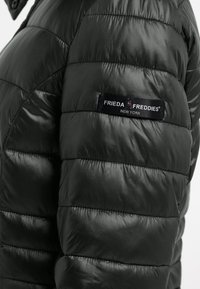 Zwarte pufferjack met een gequilte ontwerp, een gladde textuur en een merkpatch op de mouw met "FRIEDA FREDDIES NEW YORK."
