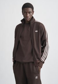 Νεαρός άνδρας που φοράει καφέ φόρμα Adidas με ροζ ανοιχτές ρίγες, στέκεται με τα χέρια στις τσέπες σε γκρι μονόχρωμο φόντο.
