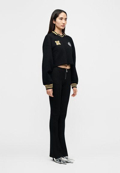 Μαύρο cropped φούτερ με κίτρινες ρίγες, κεντημένα σήματα, σε συνδυασμό με εφαρμοστά μαύρα παντελόνια και ασημί γόβες με μυτερή μύτη.