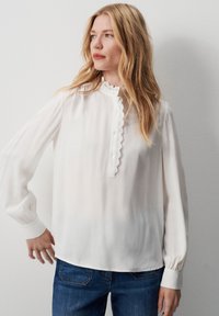 Blusa bianca a maniche lunghe con abbottonatura, colletto scallop e tessuto texturizzato. Indossata con jeans blu, caratterizzati da polsini aderenti.