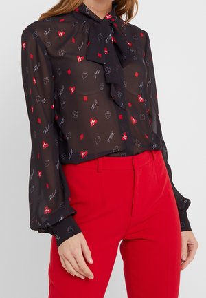 Schwarze transparente Bluse mit roten und weißen Spielkartensymbolen, gebundener Schleifenkragen, kombiniert mit hoch taillierten leuchtend roten Hosen bei einer stehenden Frau.