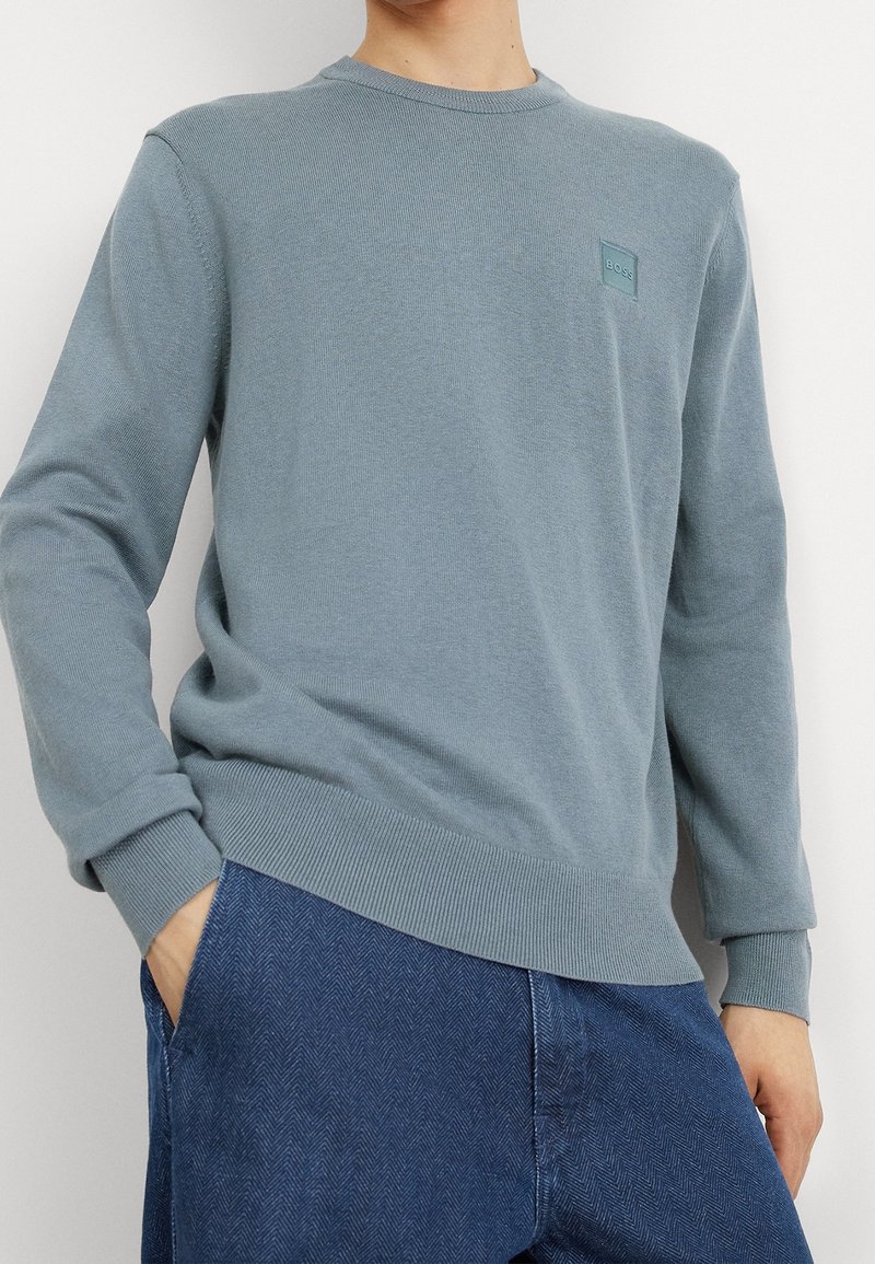 Homme portant un pull ras du cou bleu clair avec un petit logo "BOSS" sur la poitrine et un jean bleu foncé, les mains dans les poches.