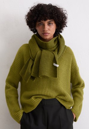 Maglione a maglia verde oliva con un motivo testurizzato, abbinato a una sciarpa coordinata. Girocollo e vestibilità comoda, indossato su pantaloni neri.