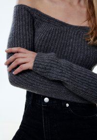 Grauer, schulterfreier Strickpullover mit gerippter Textur und langen, schmalen Ärmeln, kombiniert mit schwarzen, hoch taillierten Jeans mit Knopfverschluss.