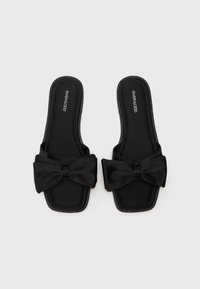 Sandale negre tip slide, fabricate dintr-un material moale, cu un detaliu mare sub formă de fundiță în față. Prezintă o talpă plată și o formă rectangulară.